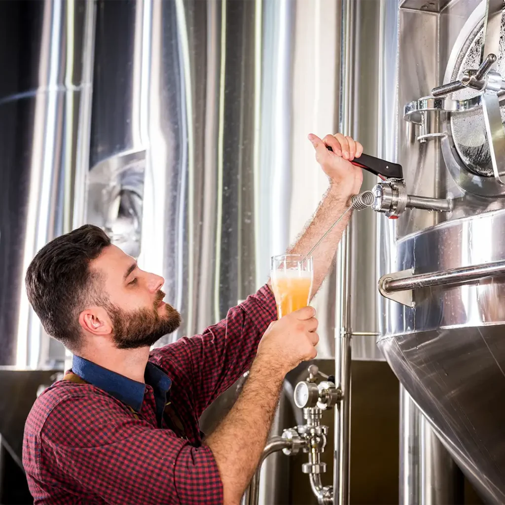 Brewer in a red plaid shirt fills a glass of beer from a stainless steel keg tap in a brewery.