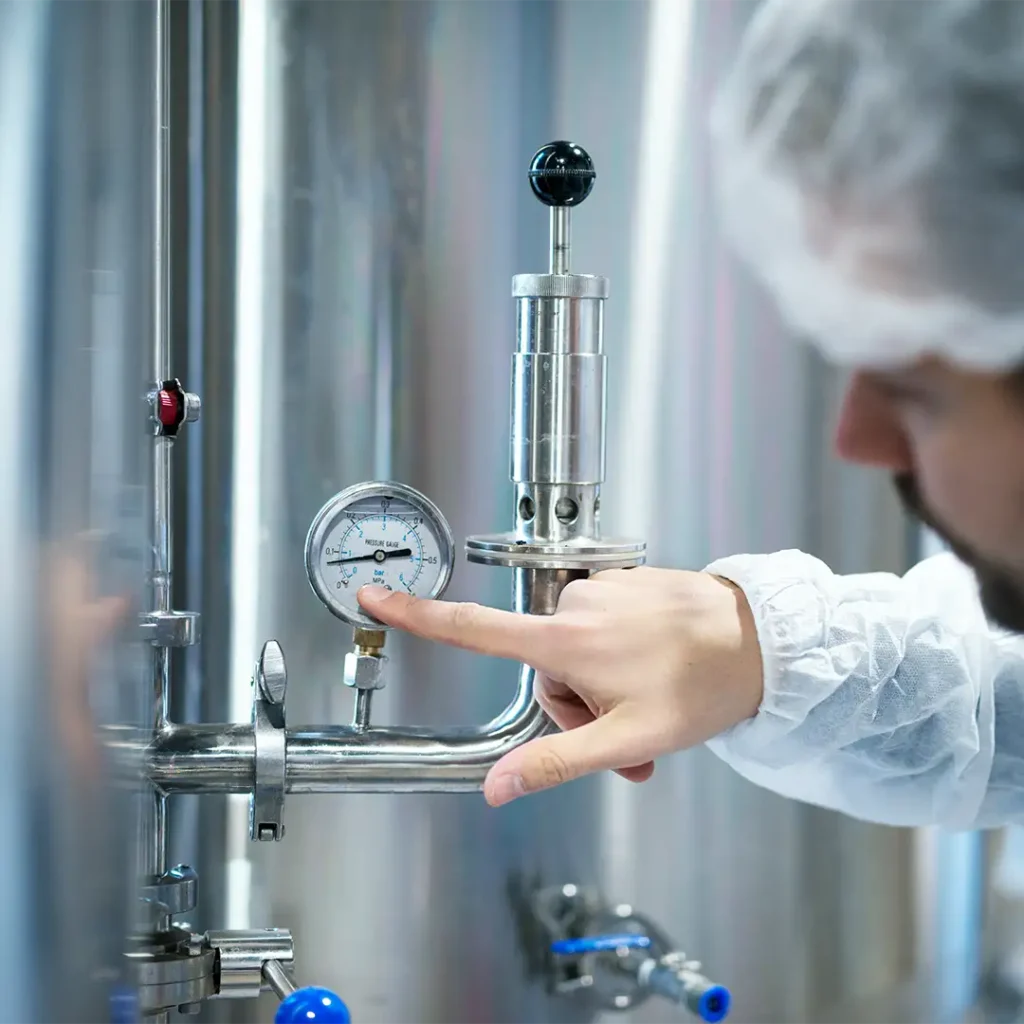 Lab worker in protective clothing adjusts a pressure gauge on stainless steel industrial equipment in a lab setting.