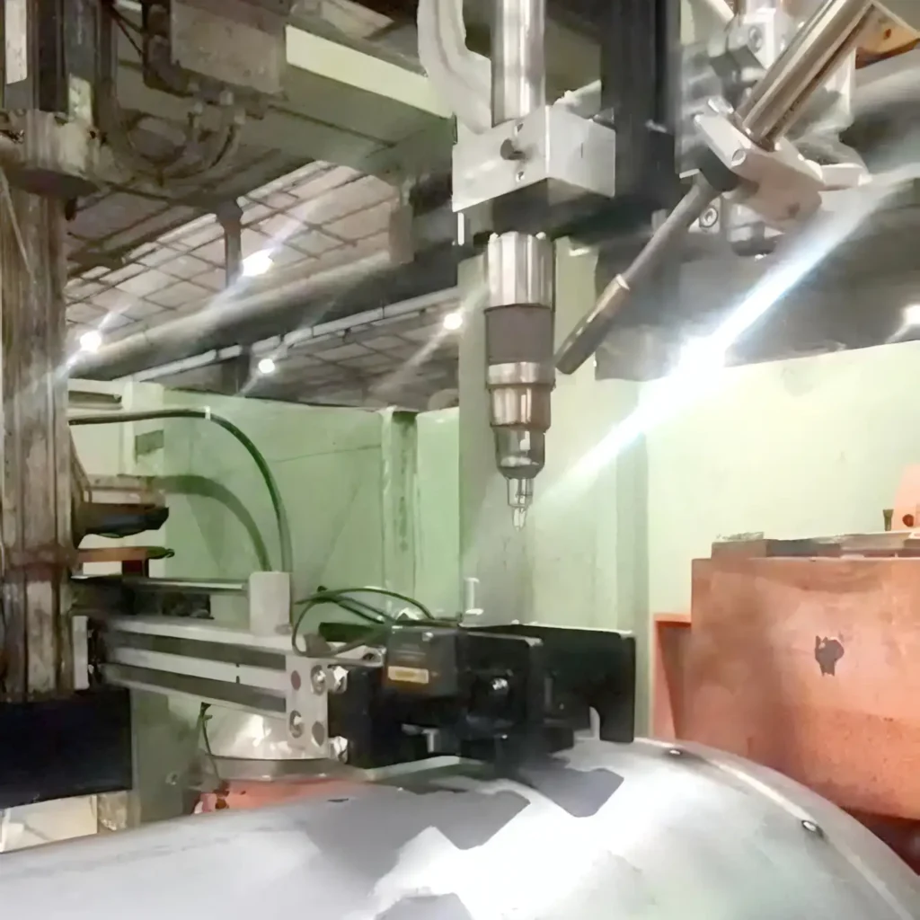 Robotic drilling spindle hanging above a metallic workpiece in an industrial CNC workshop, with green machinery and hoses nearby.